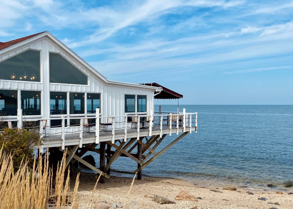 White waterfront lodge with a deck over the water and a sandy beach in Greenport, New York.