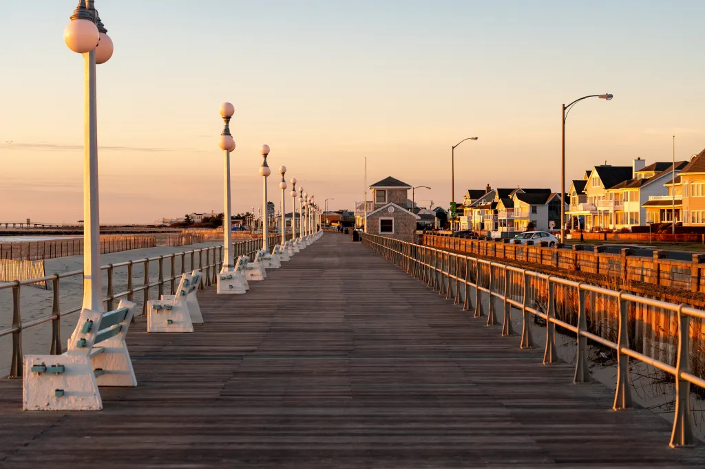 A wooden boardwalk with lampposts and benches under a golden hour sunrise in Avon By The Sea, New Jersey.