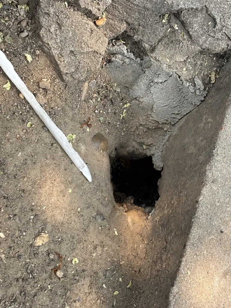 A white stick rests on brown dirt next to a dark hole in a concrete patch.
