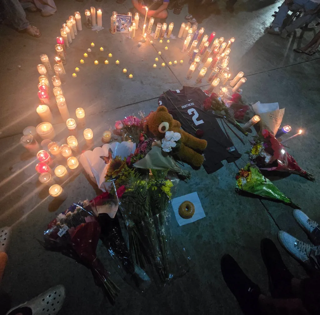 Candles arranged in a heart shape on the ground, surrounded by flowers, a teddy bear, a football jersey, and a donut.