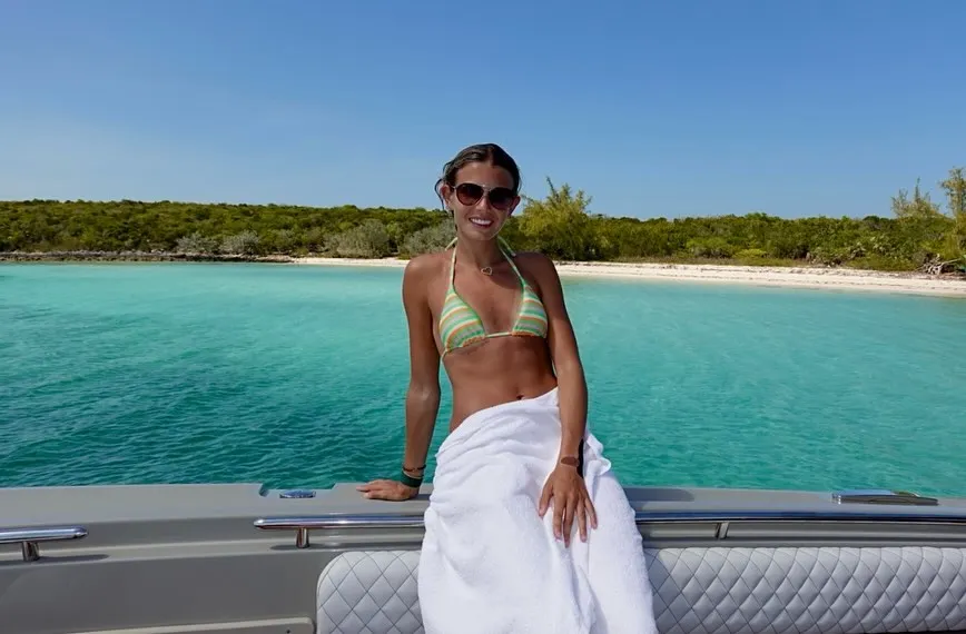 A woman in a striped bikini top and a white towel sitting on a boat with a turquoise sea and lush island in the background.