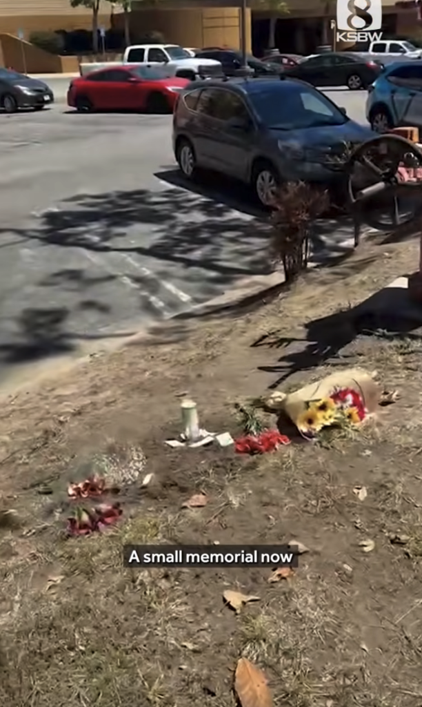A small memorial with flowers and candles on a patch of dirt next to a parking lot.