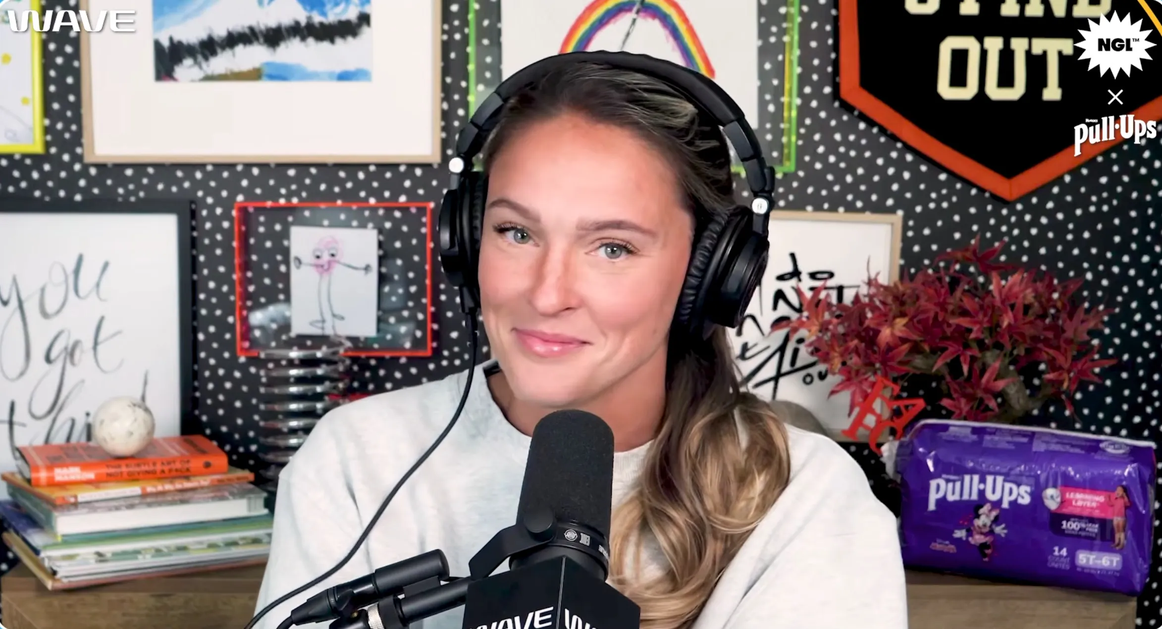 A woman with headphones and a microphone smiling in a studio with a black and white polka dot background and a pack of Pull-Ups diapers.