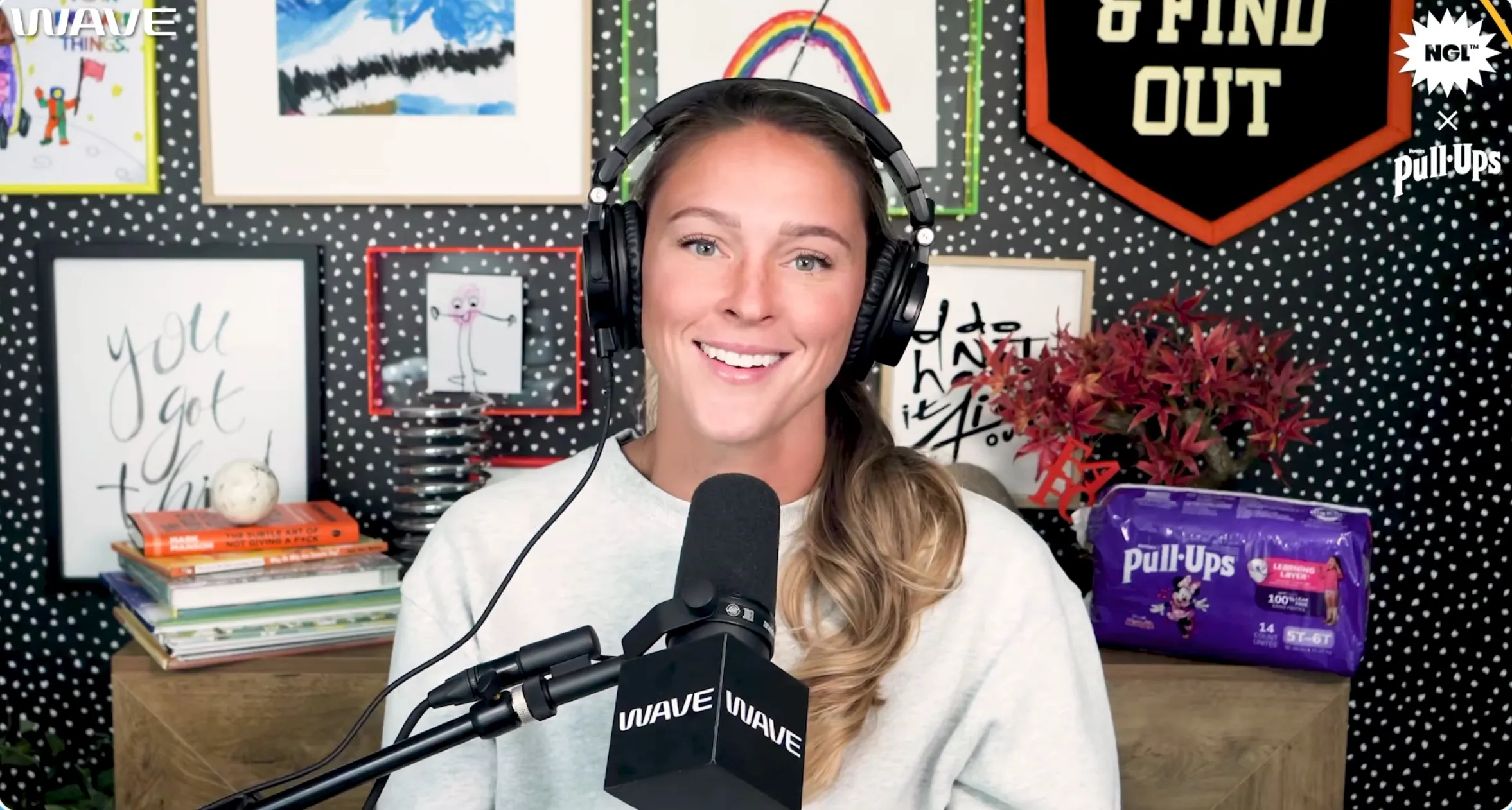 Woman wearing headphones smiles at the camera, a package of Pull-Ups diapers to her right.