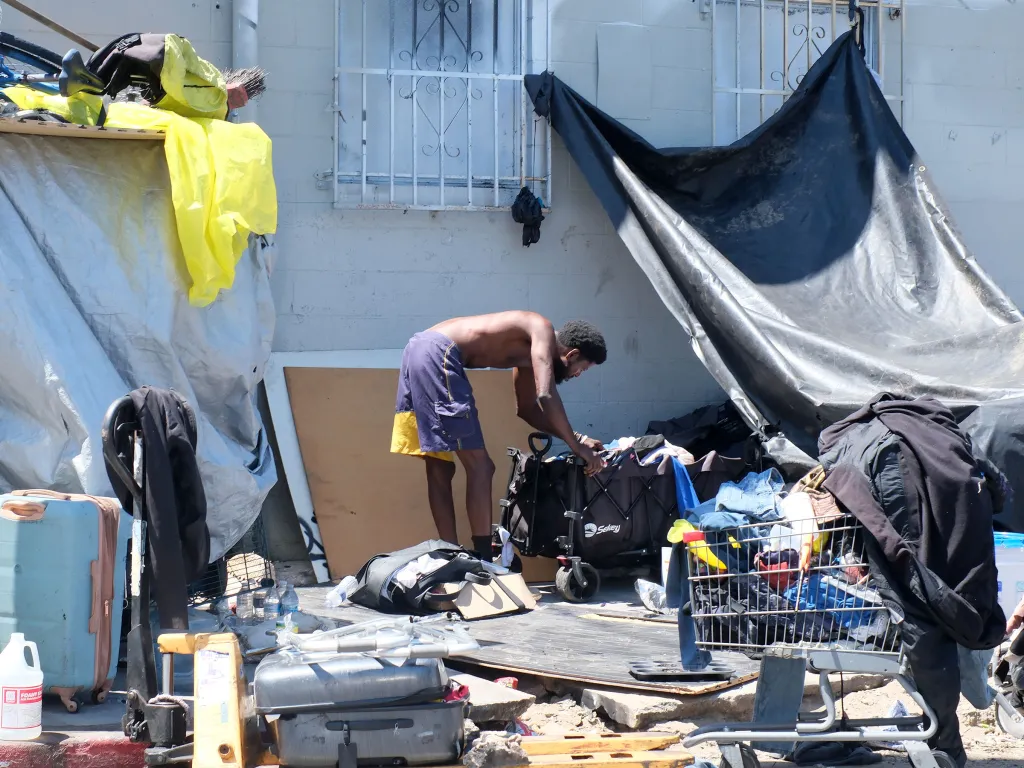 A shirtless man bent over a black pushcart filled with belongings next to a tarp-covered structure on the street.