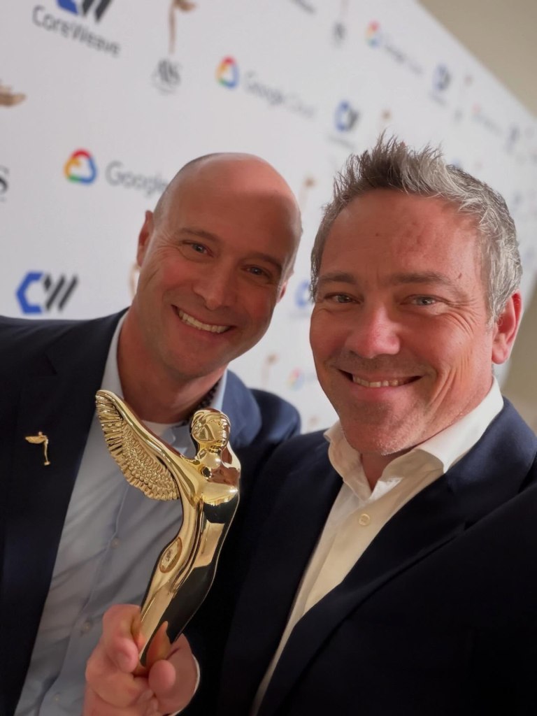 Two men smiling for a selfie, holding a golden winged trophy.