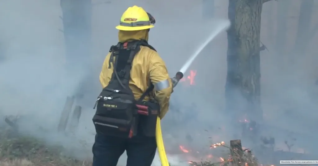 A firefighter in yellow gear aims a hose at a fire in a smoky, wooded area.