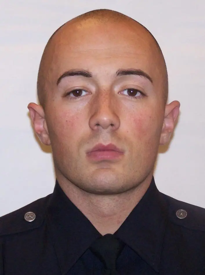 Headshot of a man with shaved head wearing a dark uniform shirt and tie.
