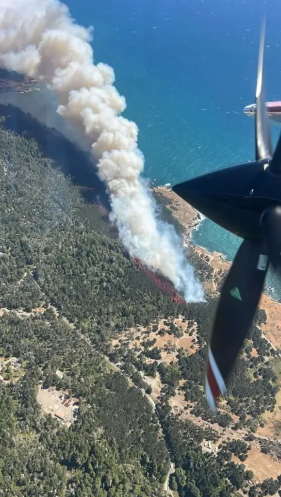 Aerial view from a plane of the Woodside Fire in Sonoma County, showing a long plume of smoke rising from a forested area along the coast.
