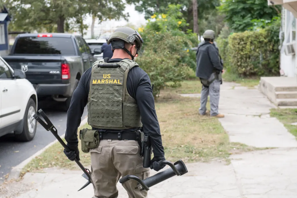 A U.S. Marshal in tactical gear, carrying tools, stands outside a house during Operation Triple Beam in San Antonio.