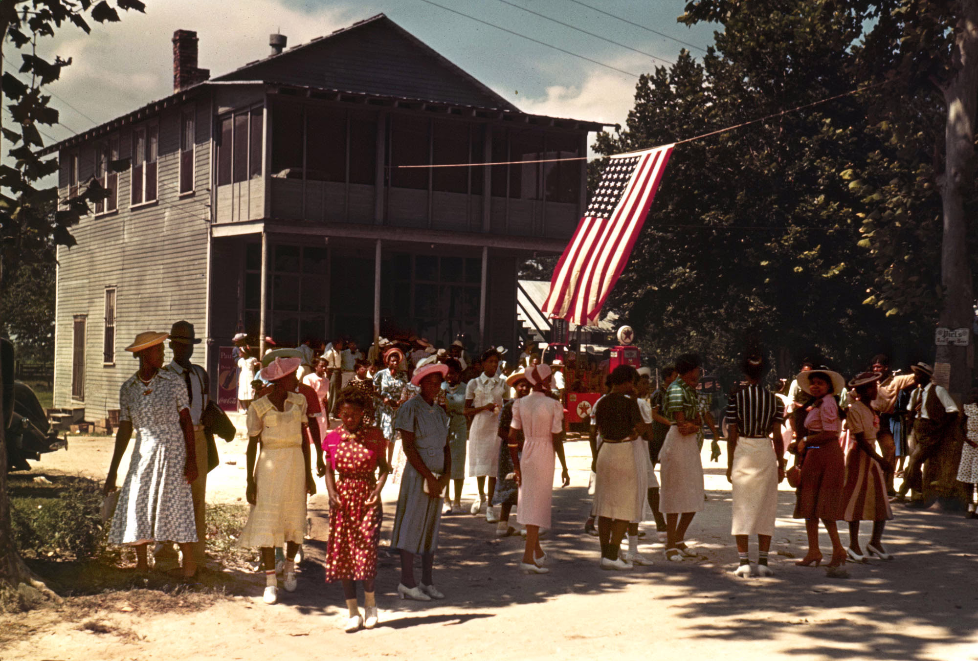 Dozens of well-dressed people stand along a road, beside a Texaco station, on the Fourth of July in 1939.