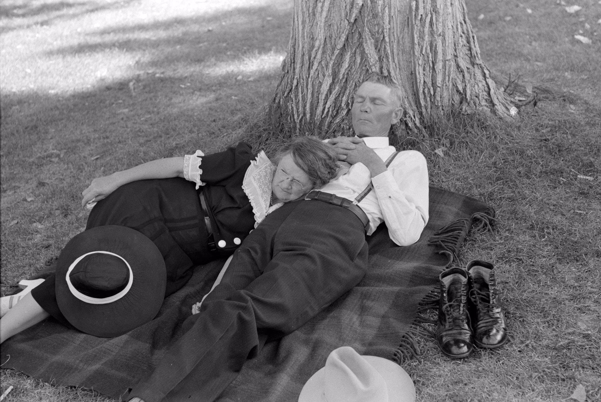 An older couple lies napping together on a blanket at the shady base of a tree.