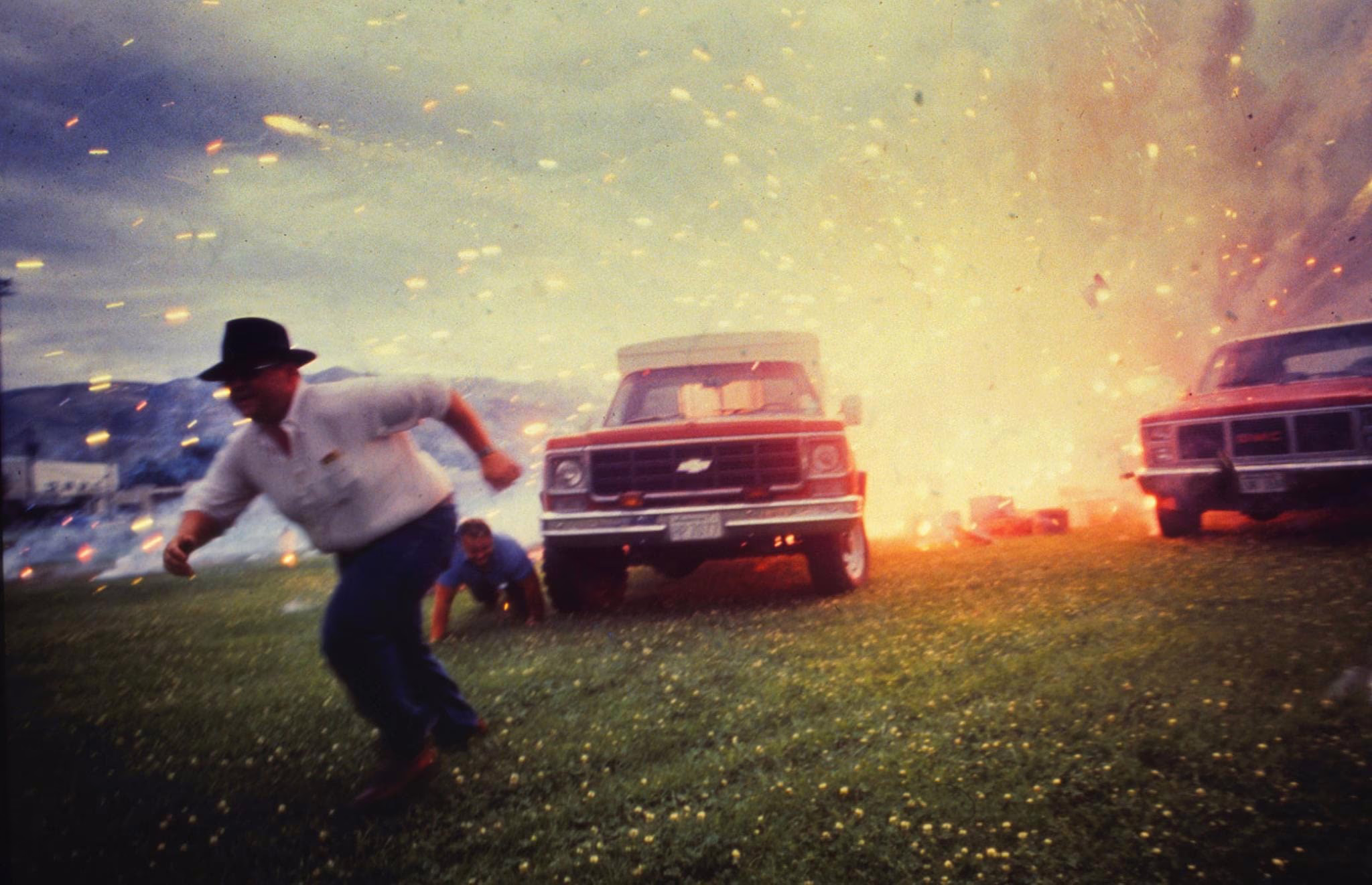 Two people scramble, running past trucks as fireworks light up close behind them.