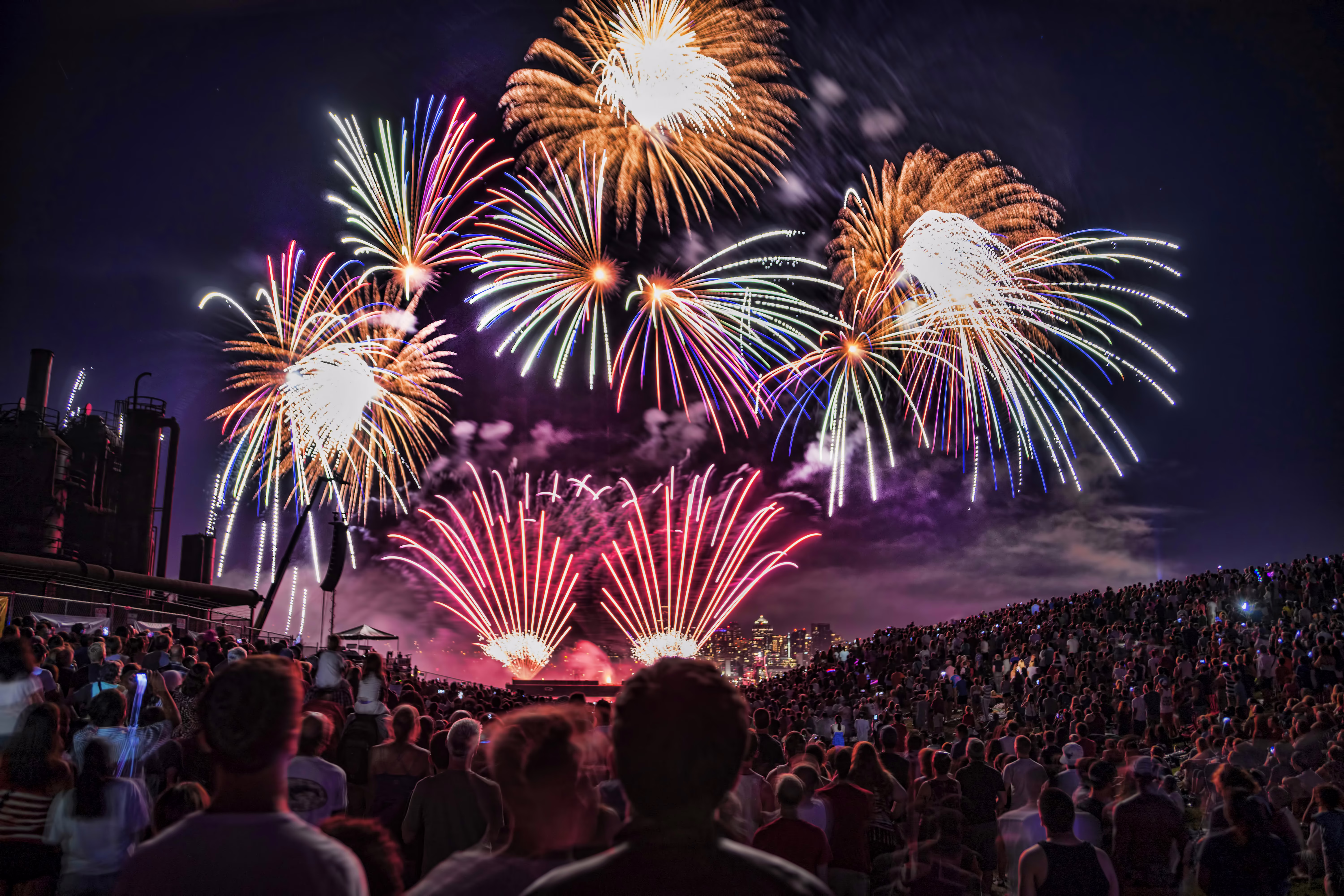 A crowd in a park enjoys Fourth of July fireworks, with Seattle's skyline in the background.