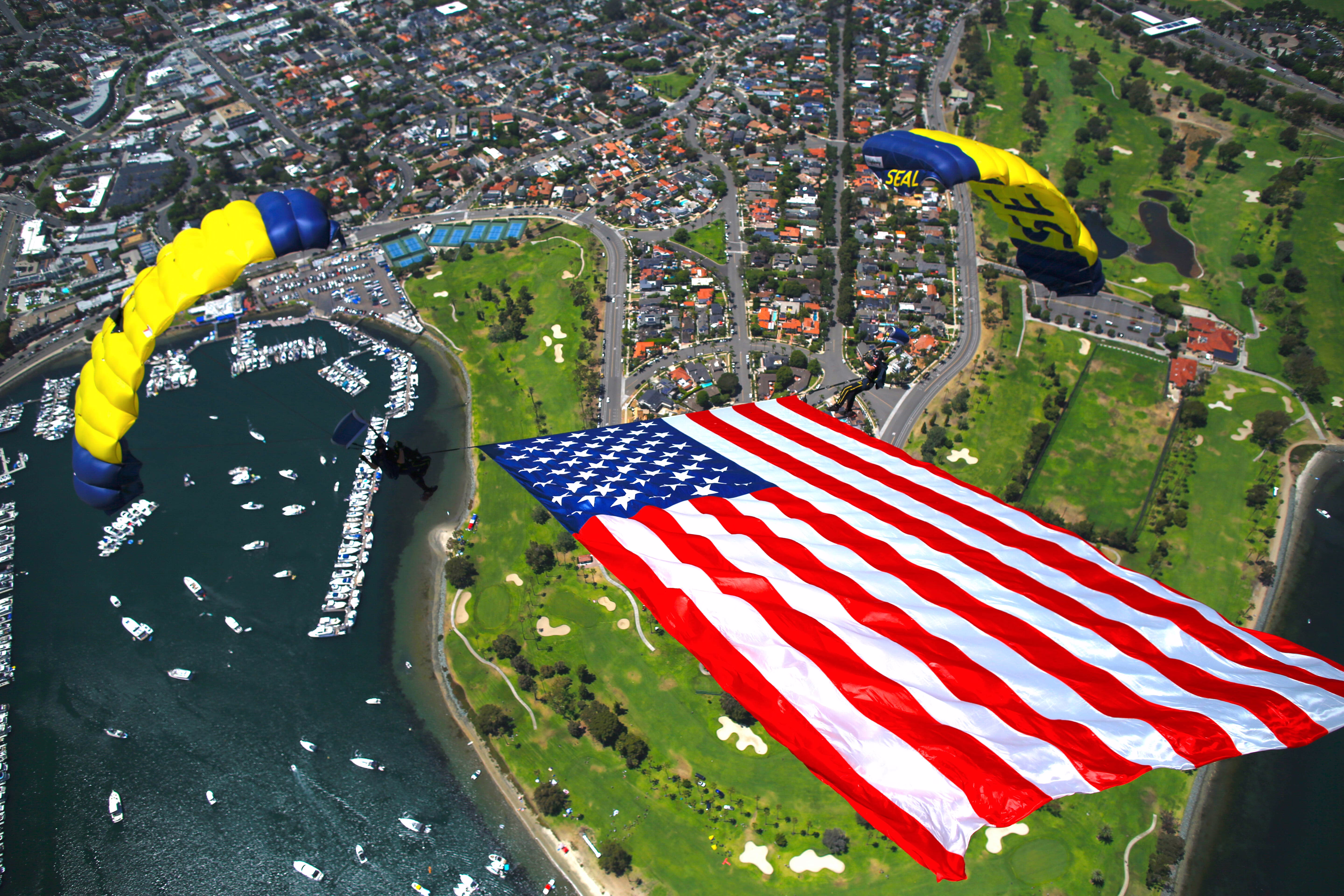 An aerial view of two Navy parachutists towing a large American flag tethered between them, above a small-boat harbor and a golf course.