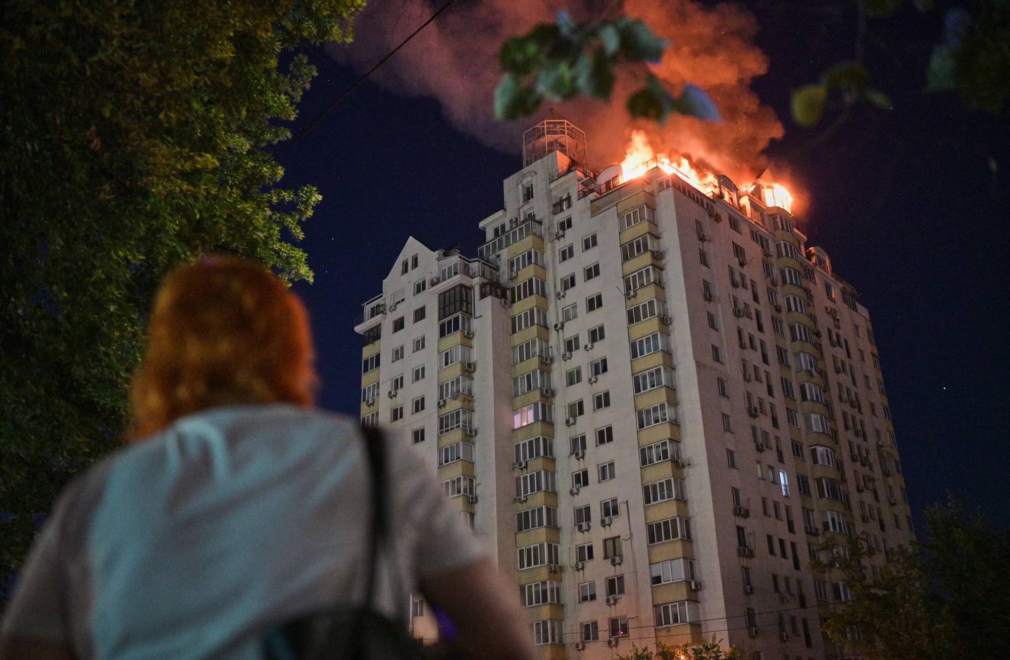 A woman looks up at a tall burning apartment building at night.