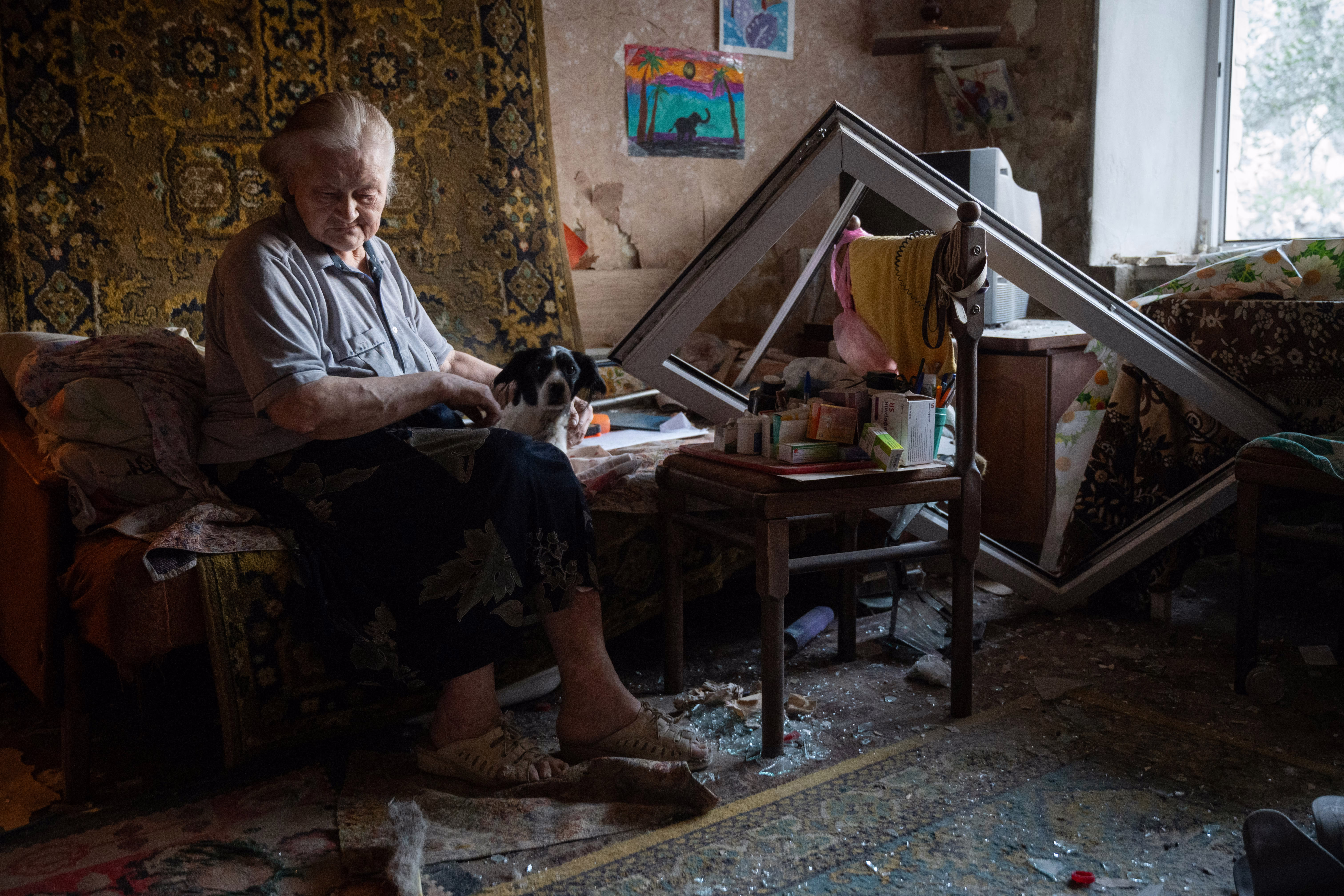 An older woman sits in a room in an apartment, surrounded by debris and a broken window frame, after a nearby missile strike.