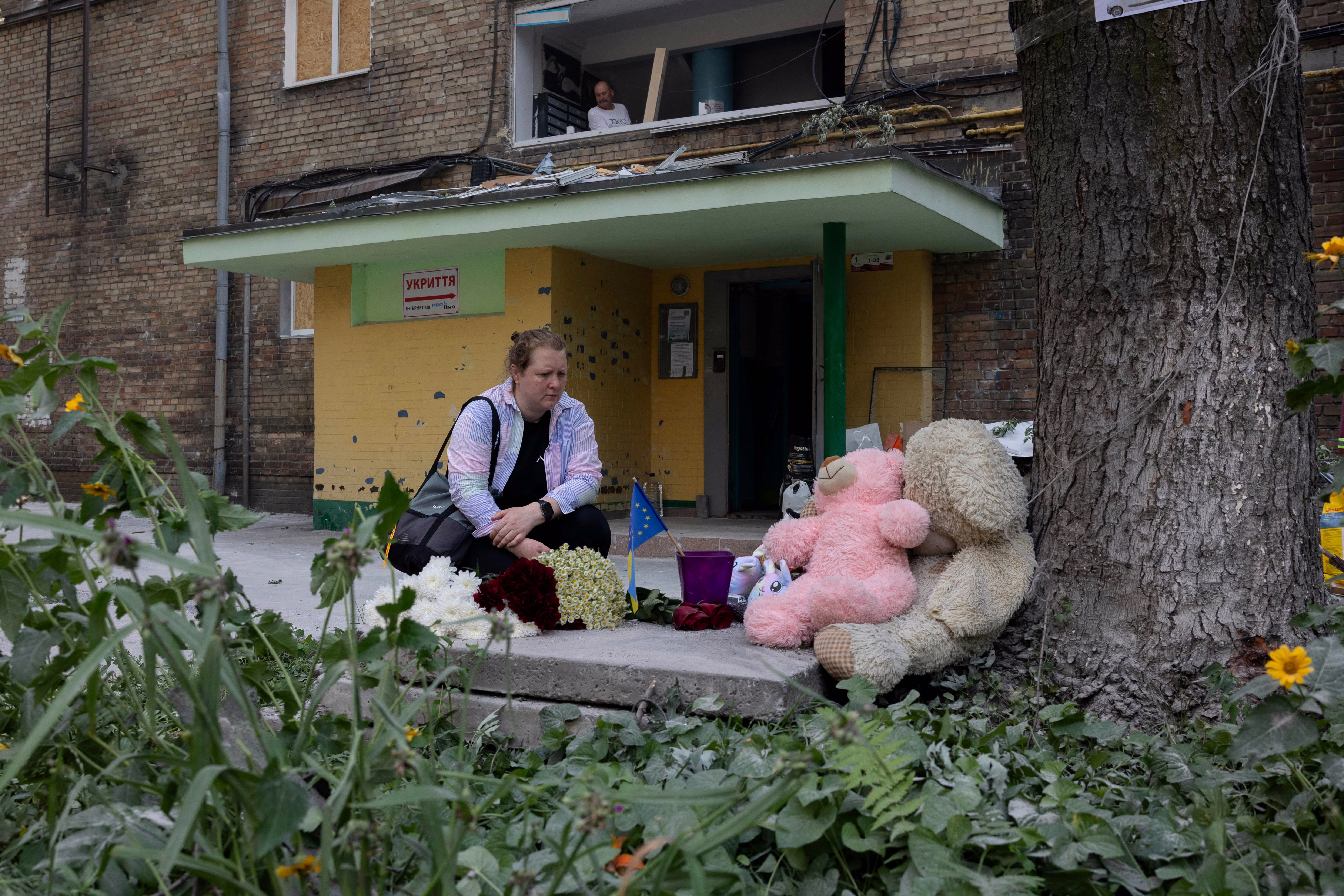 A woman kneels next to a makeshift memorial beside a damaged residential building.