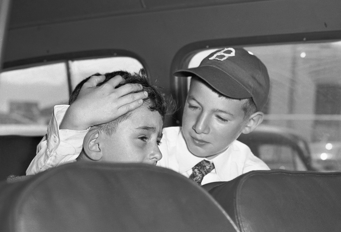 black-and-white archival photo of boy in white shirt, tie, and baseball cap comforting a younger boy crying inside a vehicle