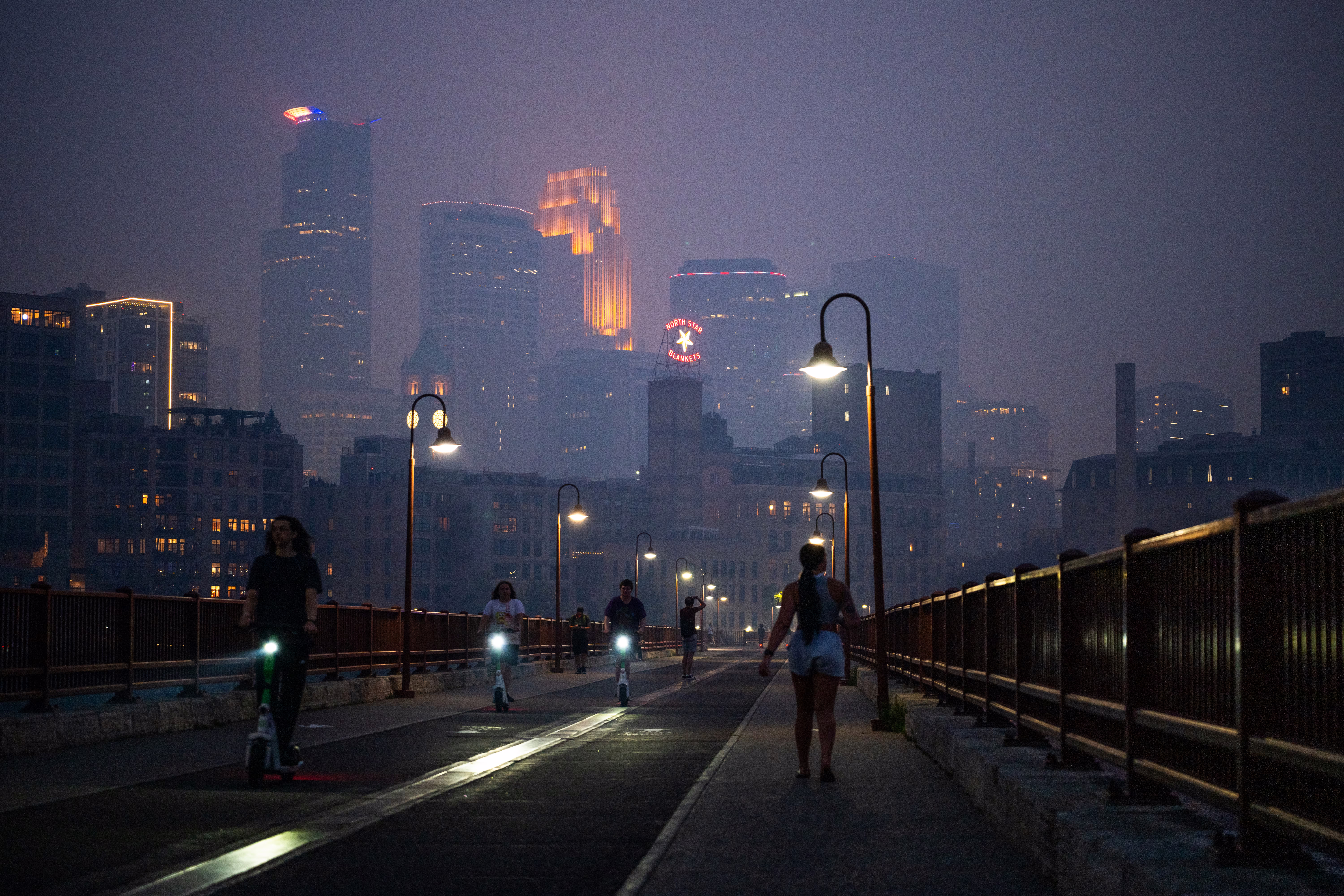 People walk on a bridge at night, with tall city buildings in the background obscured by smoke.