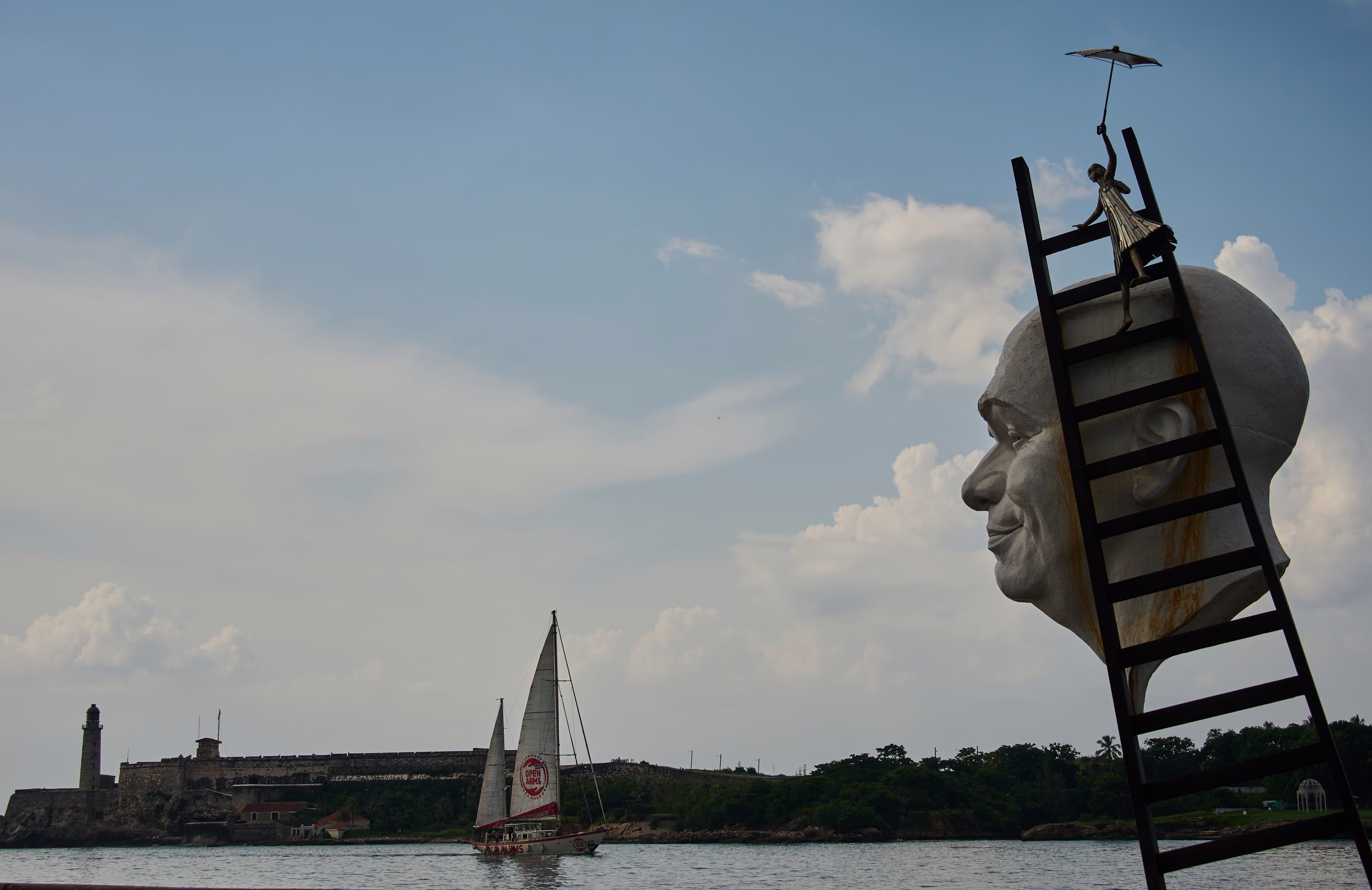 Artwork seen near a harbor, featuring a large sculpture of a man's head attached to a ladder, with an umbrella-holding girl climbing on it.