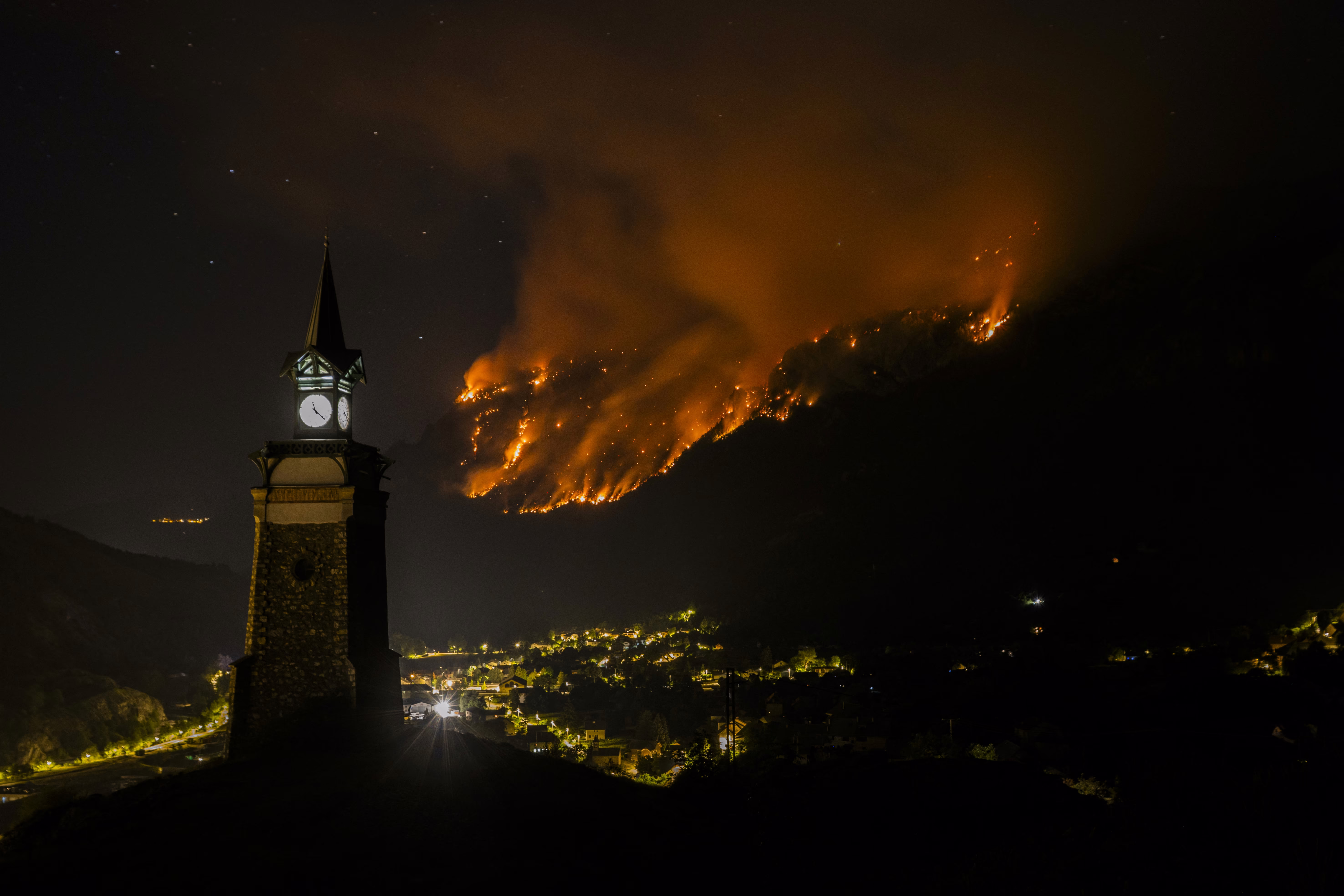 An alpine village seen at night, beneath a wildfire burning across a mountainside
