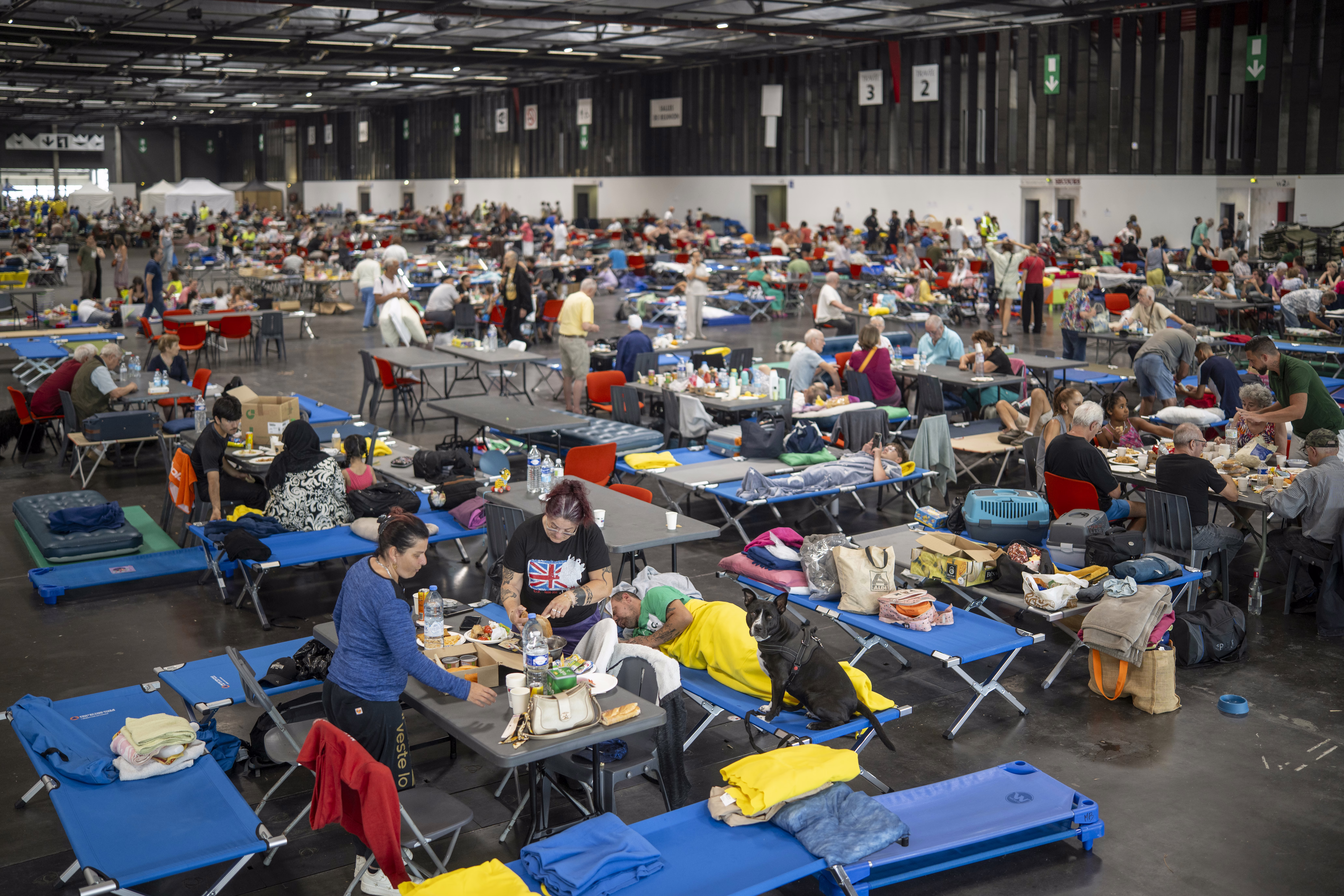People gather in a temporary shelter building, sitting at tables and on cots.