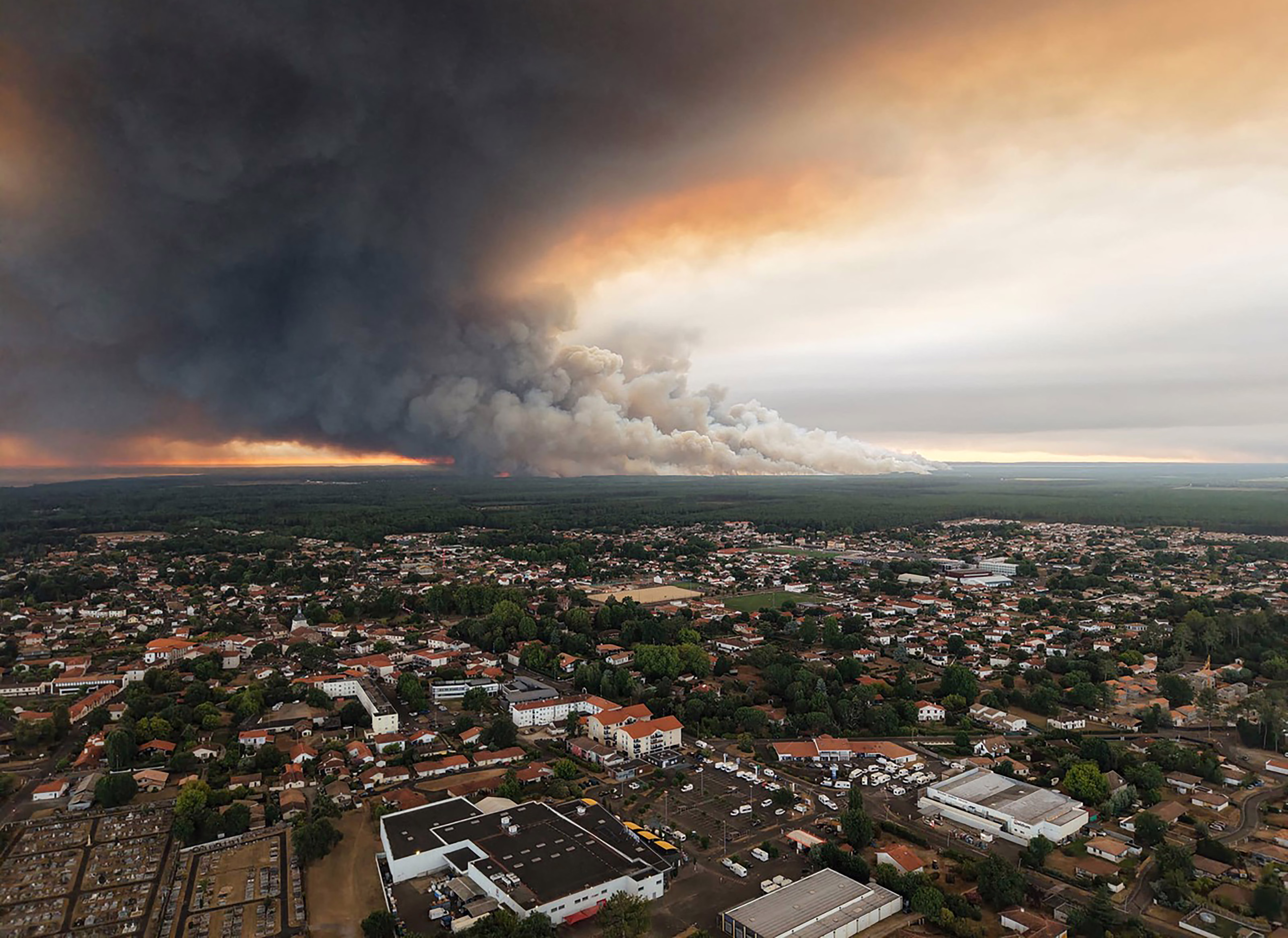An aerial view of a wildfire spreading toward a city, sending huge plumes of smoke into the sky