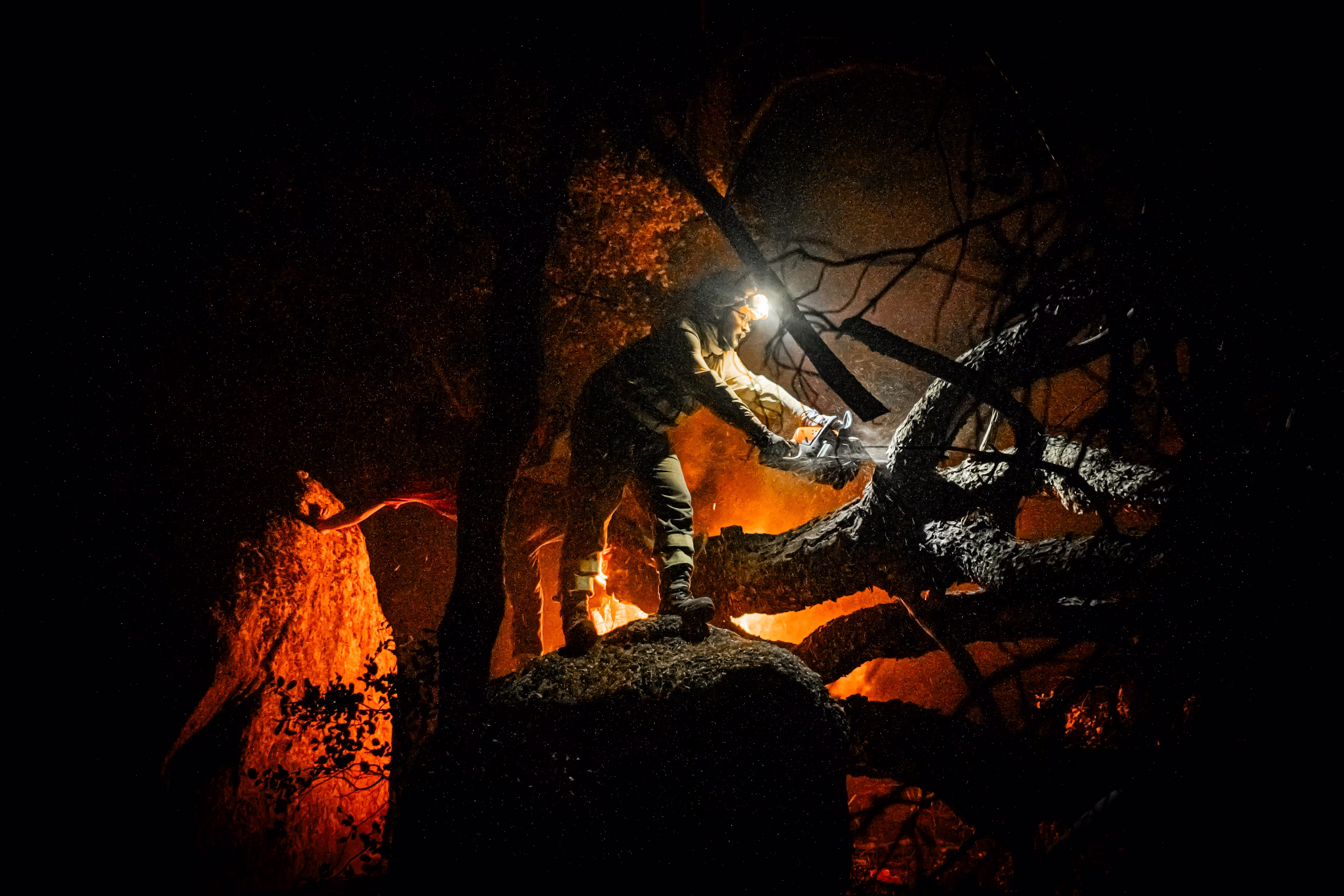 A person stands on a rock and uses a chainsaw to cut large branches from a fallen and burning tree.