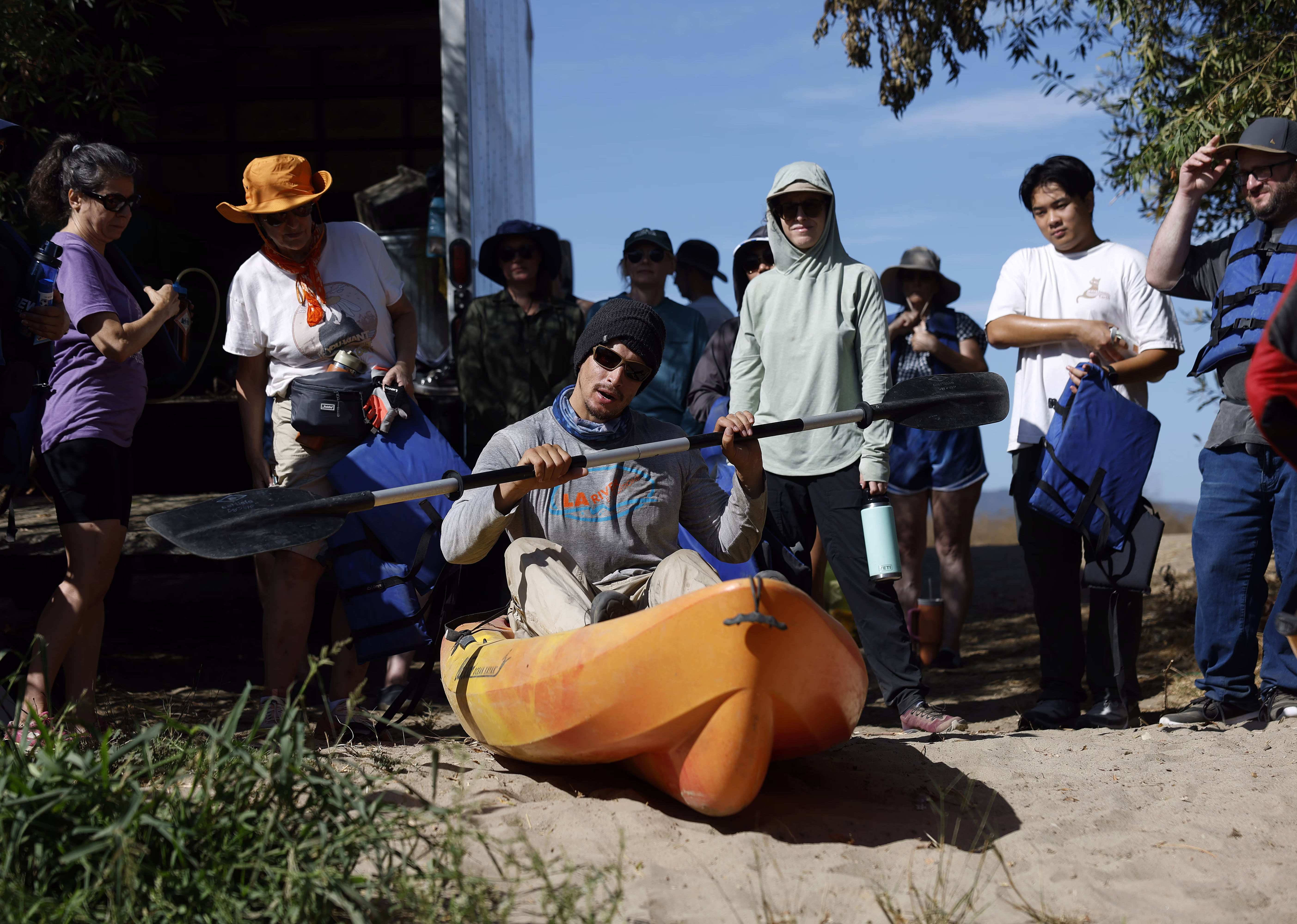 A person demonstrates how to paddle from inside a kayak on a riverbank, as a group watches.