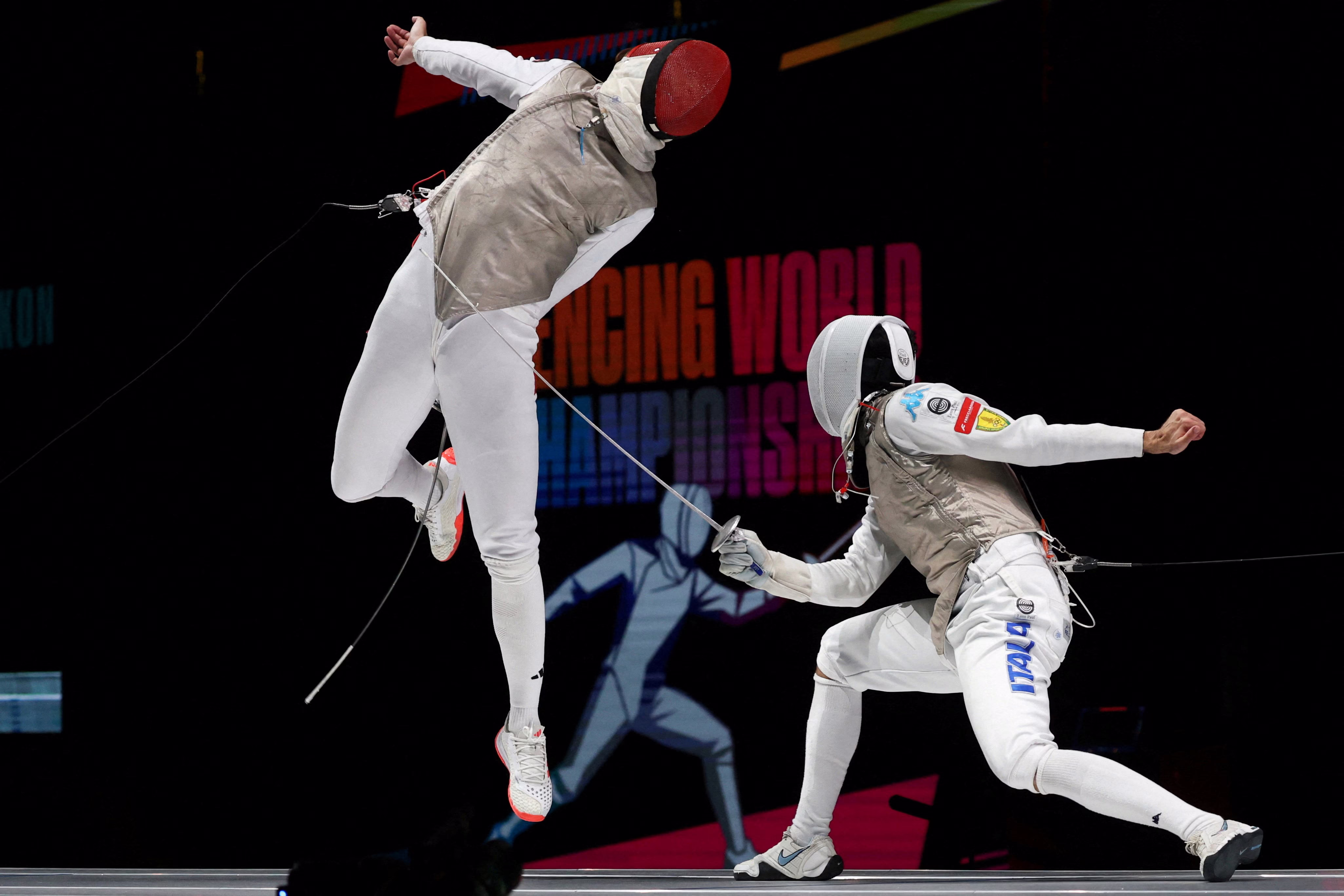 Two fencers duel in a competition, as one leaps high to evade an attack.