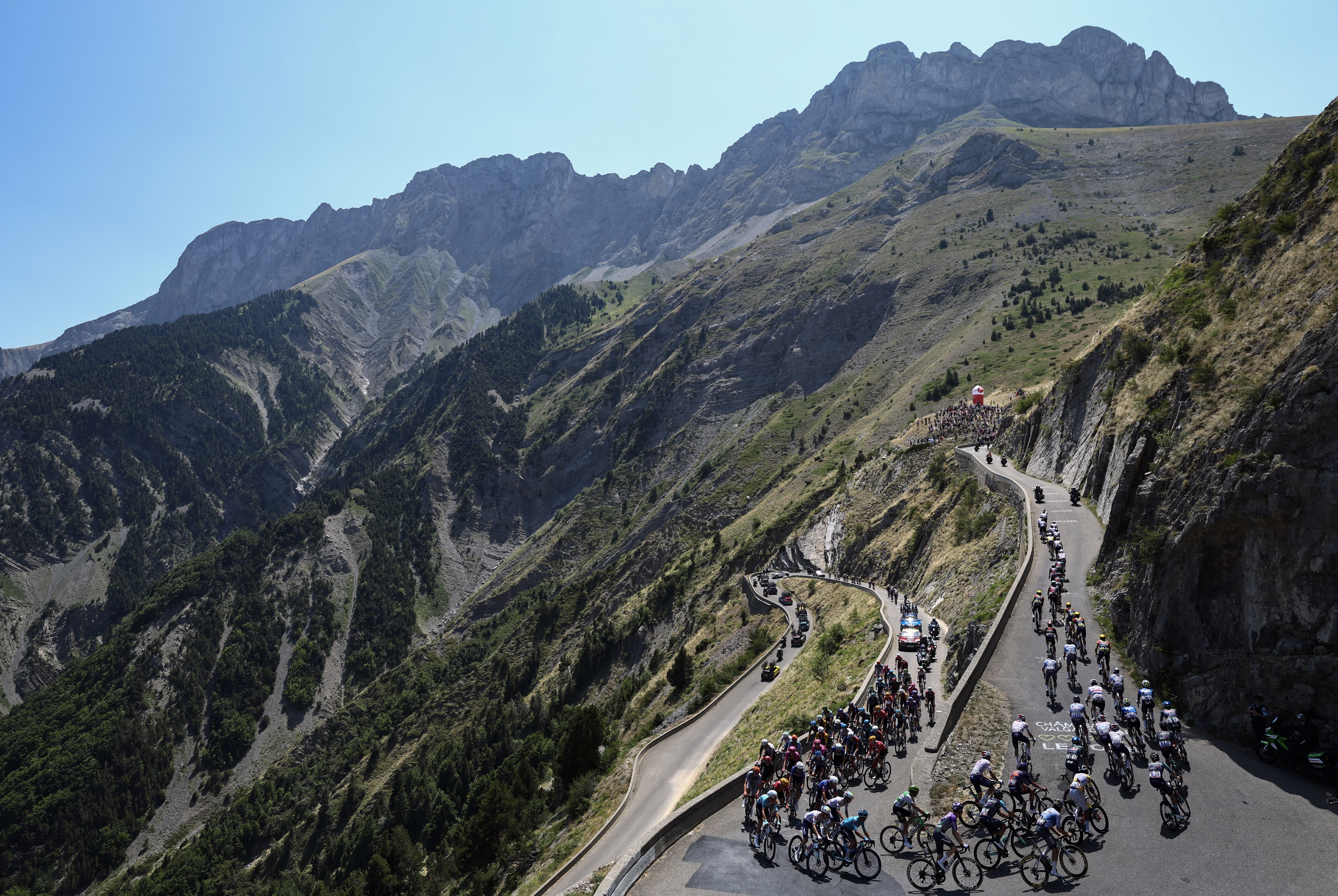 A pack of cyclists rides up a switchback road on a steep mountainside.