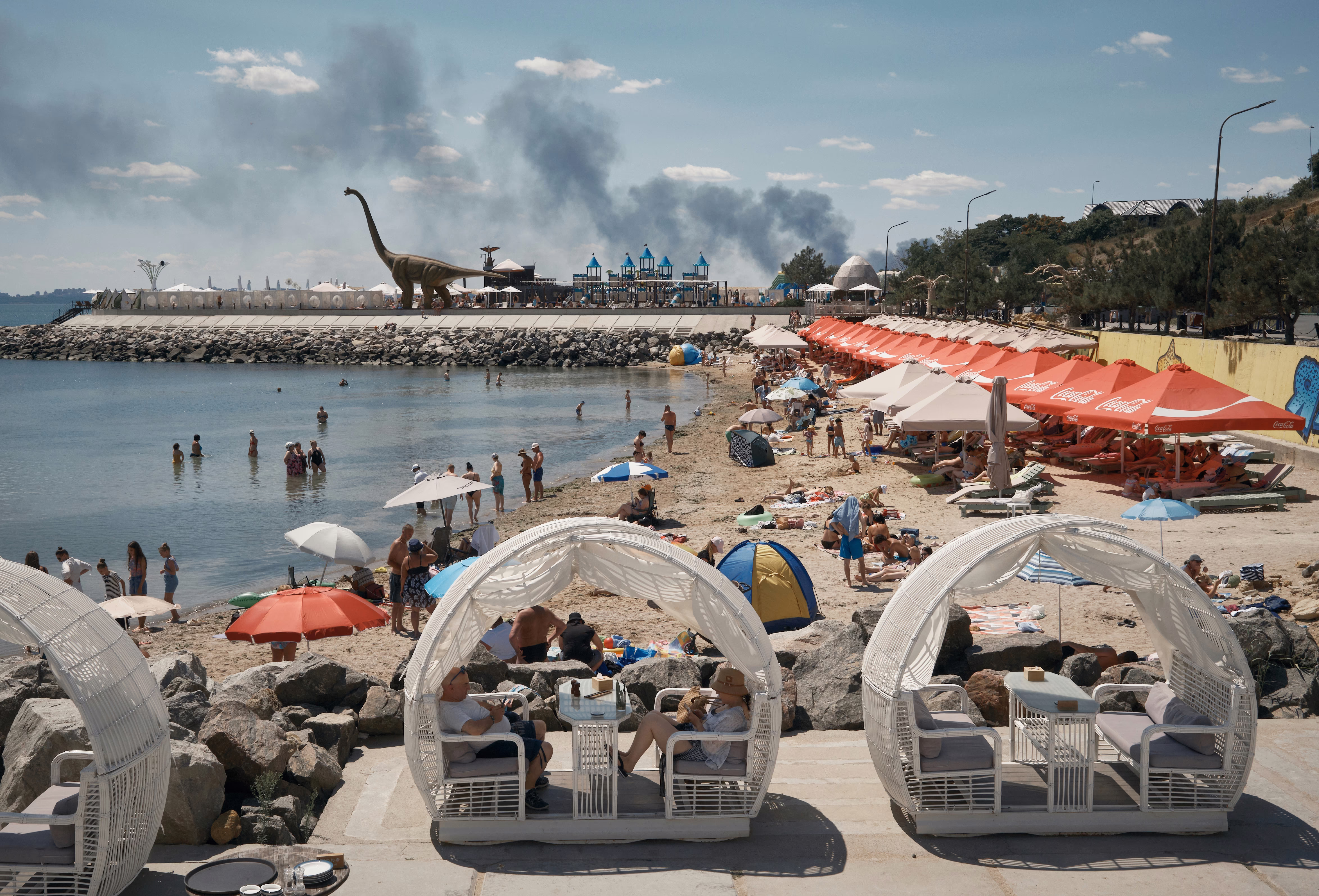 Smoke rises in the background, beyond a beach resort area, featuring a life-sized dinosaur statue.
