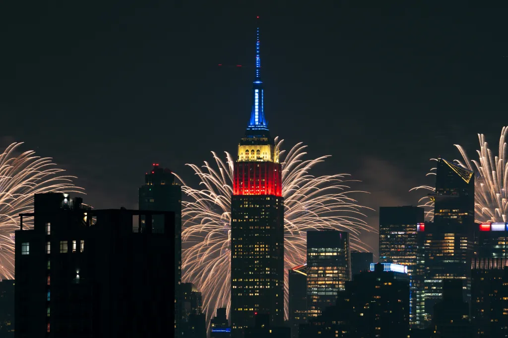 Fireworks light up the night sky behind the Empire State Building, which is illuminated in red, white, and blue.