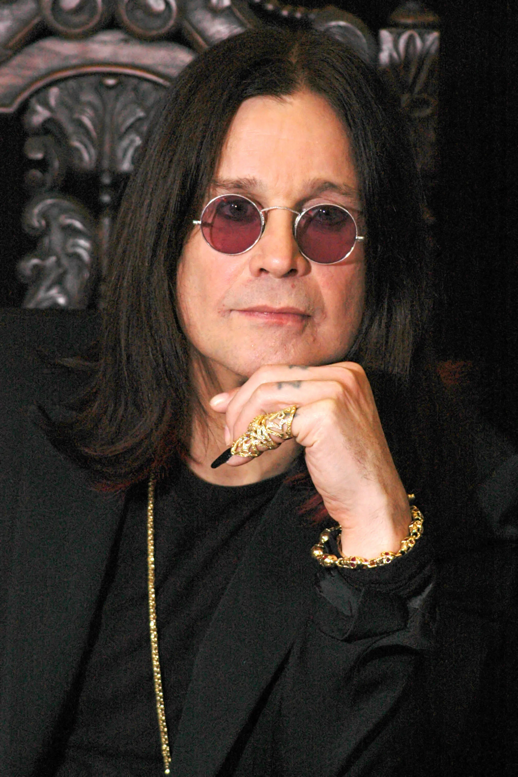 Ozzy Osbourne, wearing red-tinted round glasses, a black suit, and a long gold necklace, poses with his chin resting on his right hand which is adorned with a golden ring, with a darker background.