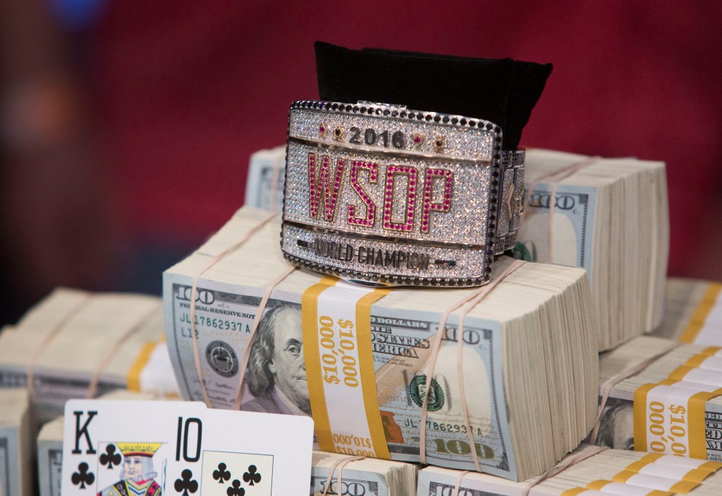 The 2016 World Series of Poker championship bracelet displayed on stacks of cash, with playing cards King and 10 of Clubs in the foreground.
