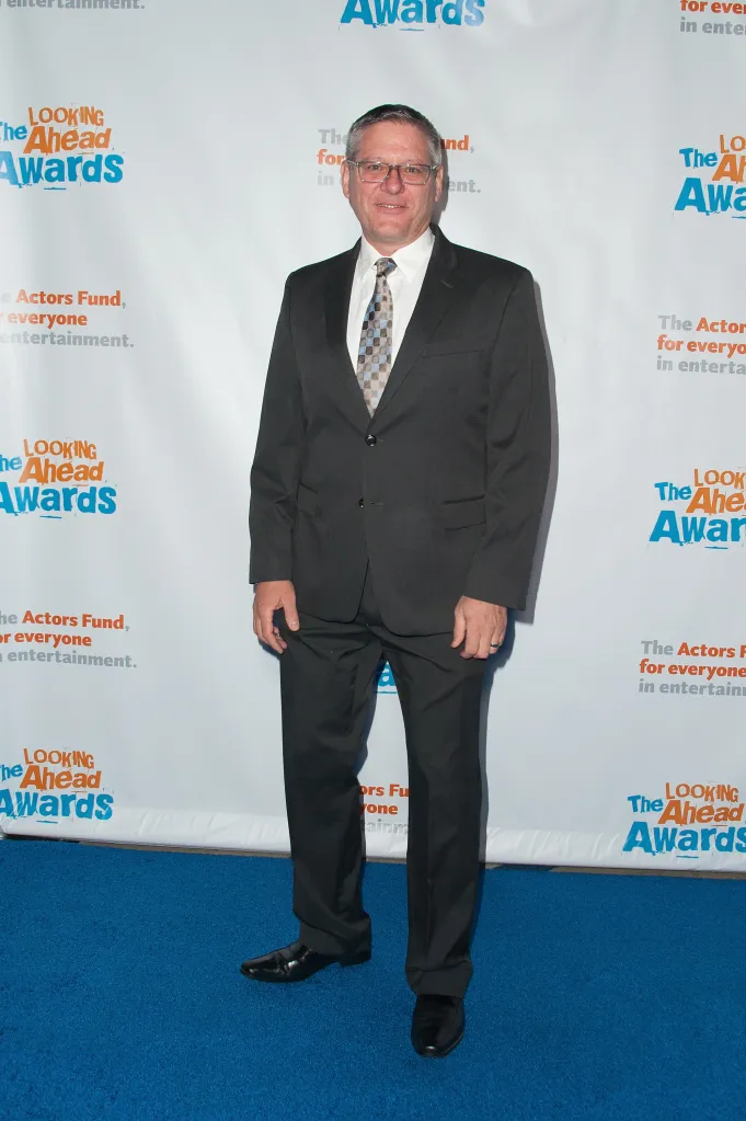 Radames Pera smiling and wearing a suit and patterned tie on a blue carpet in front of a white backdrop with "The Looking Ahead Awards" logos.