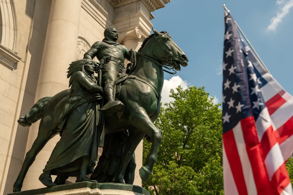 Equestrian statue of Theodore Roosevelt next to a Native American man and an African man, with an American flag in the foreground.