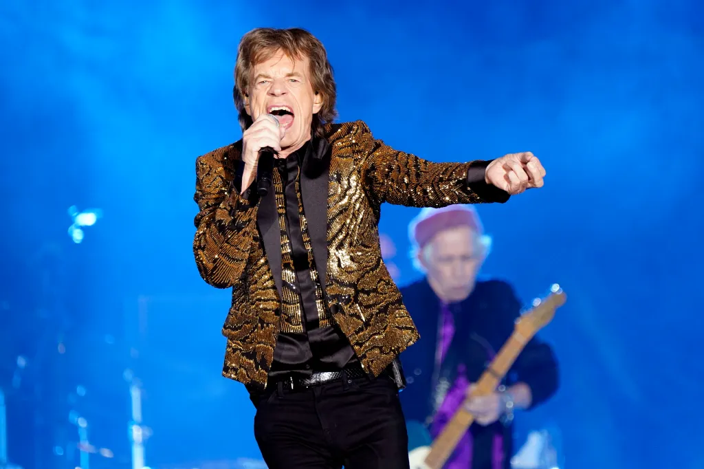 Mick Jagger of The Rolling Stones singing into a microphone on stage, with a guitarist blurred in the background.