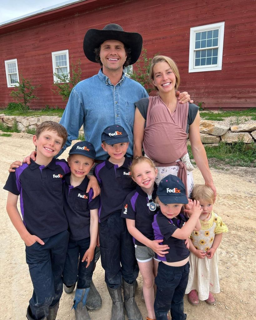 Hannah Neeleman and her husband, with their seven children, including an infant in a carrier, standing in front of a red barn.