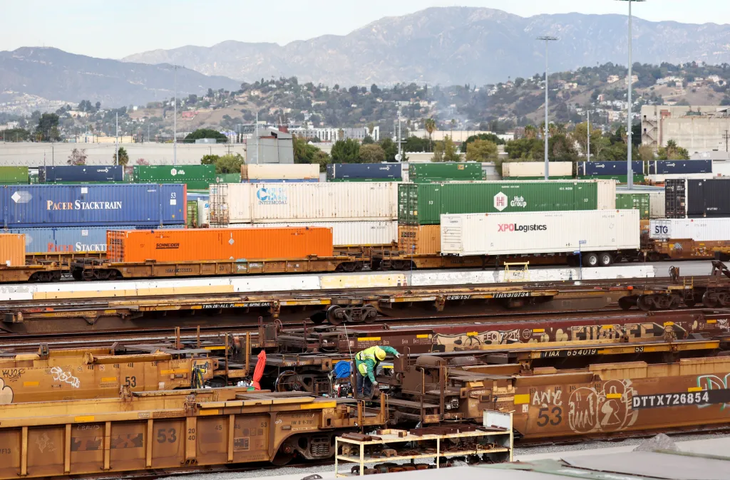 A worker in a yellow vest and hard hat works on a rail car in a Union Pacific Intermodal Terminal rail yard, with stacked shipping containers and mountains in the background.