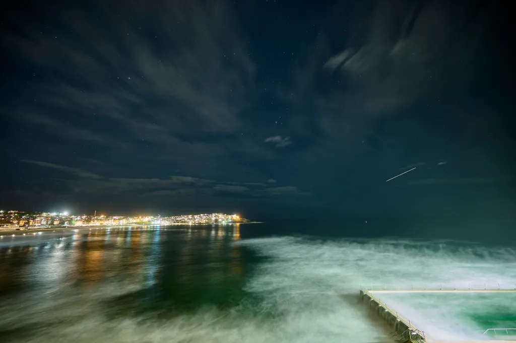 A meteor streaks across the dark night sky above a brightly lit Sydney coastline and ocean pool.