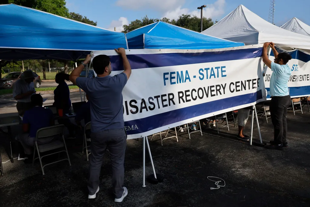 Two FEMA employees hanging a banner that says "FEMA - STATE DISASTER RECOVERY CENTER" under a blue tent.