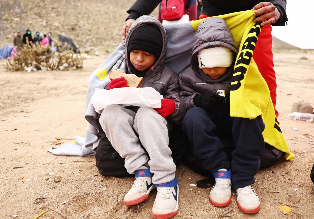Two young asylum-seeking migrants are warmed by a donated blanket, one holding a piece of bread and the other a small snack.