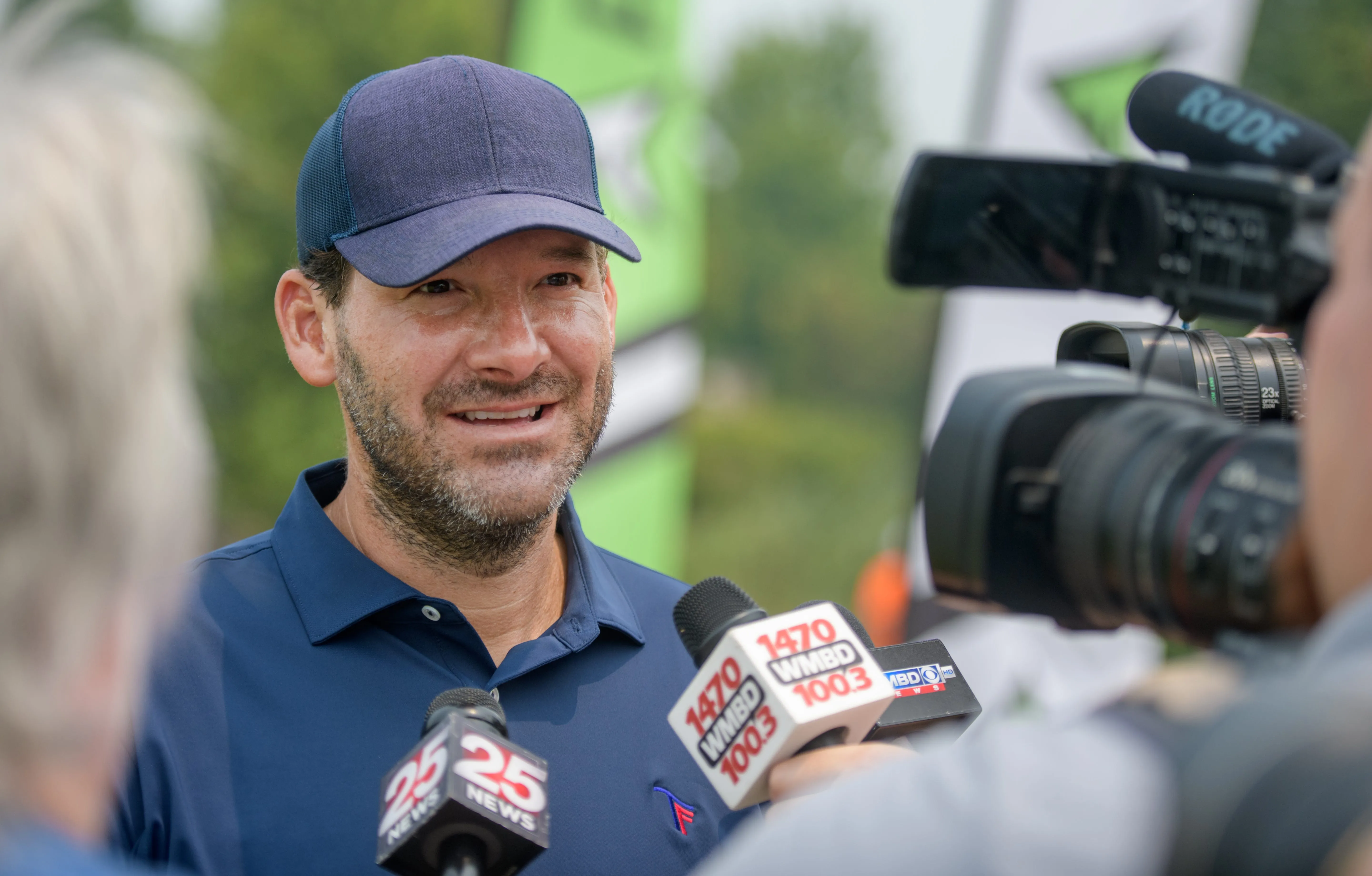 Tony Romo, wearing a blue hat and shirt, speaking into several microphones at a media event.