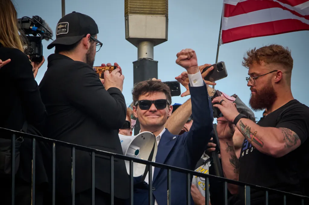 Nick Fuentes wearing sunglasses and a blue suit, holding a megaphone and raising his fist, surrounded by supporters holding phones.
