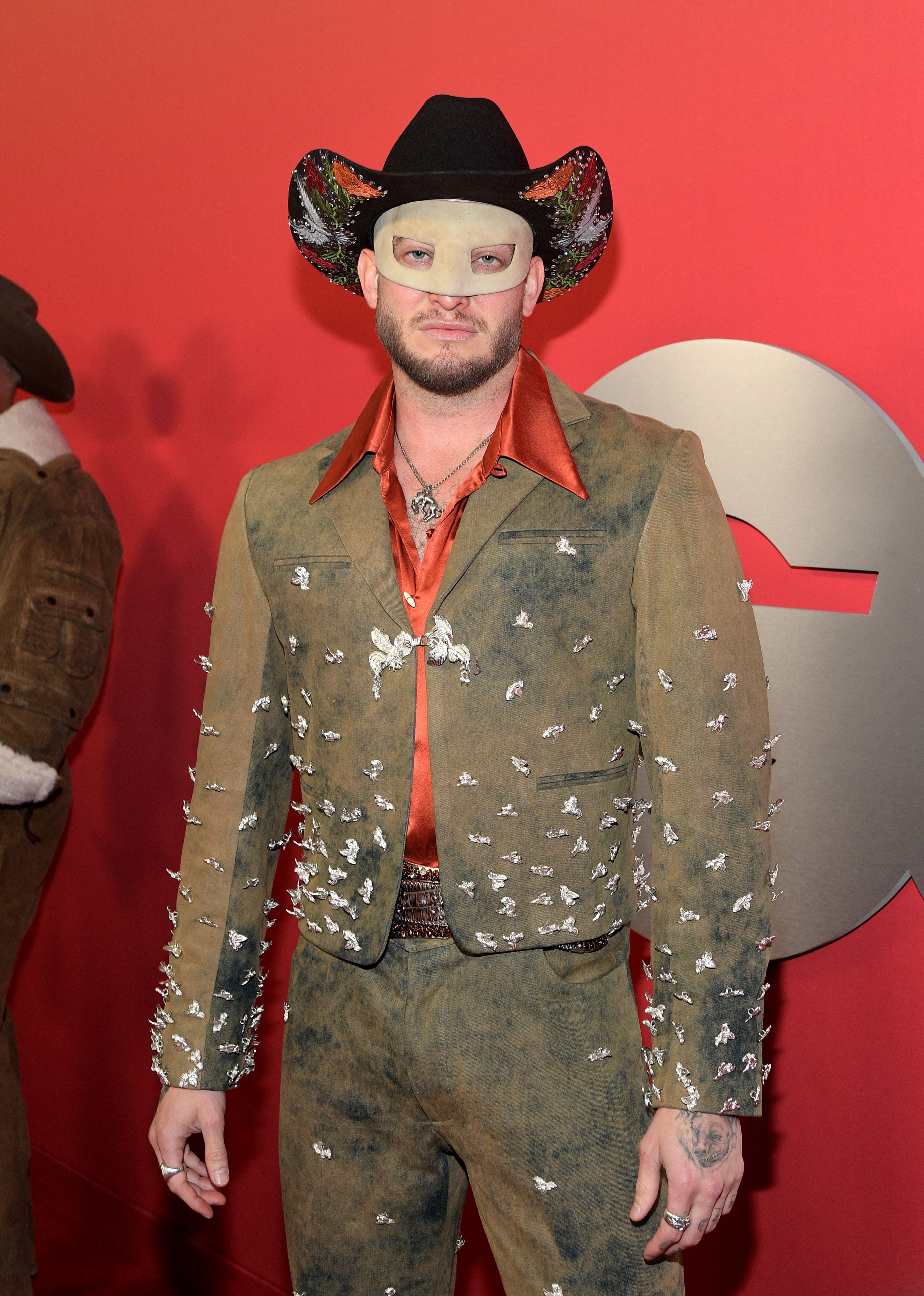 Orville Peck wearing a full brown suit adorned with silver embellishments, an orange shirt, a black cowboy hat with colorful designs, and a light-colored mask covering his eyes and nose.