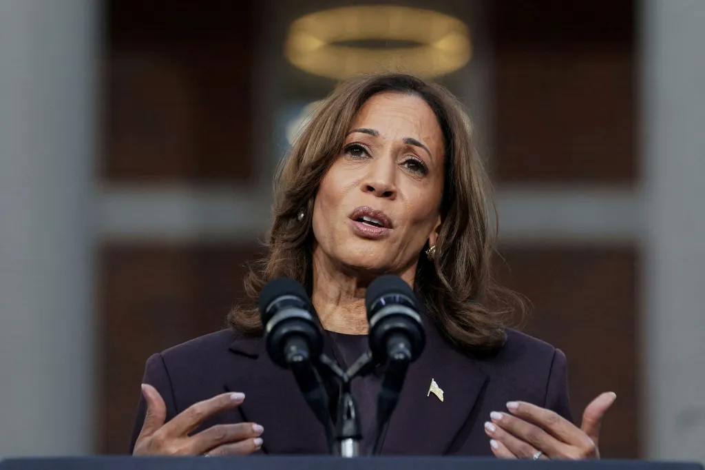 Kamala Harris speaking at a podium with two microphones, her hands gestured open.