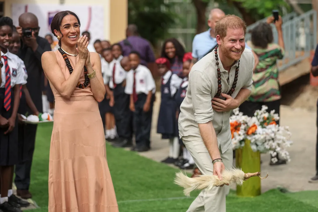 Meghan, Duchess of Sussex, clapping while Prince Harry, Duke of Sussex, smiles and bows, holding a white feathered object during an Invictus Games anniversary event.
