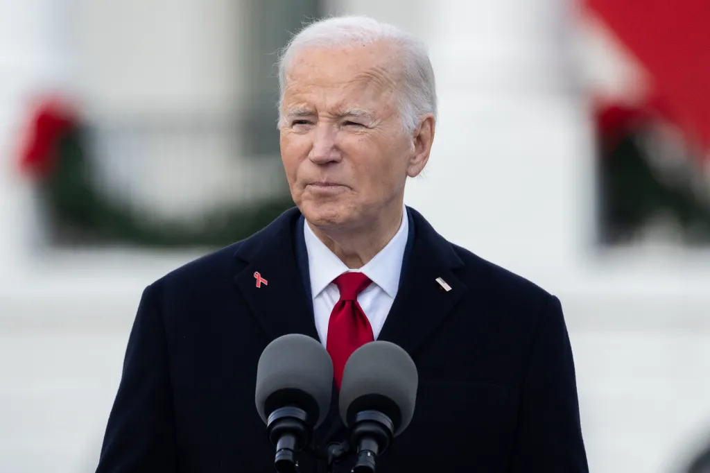 President Joe Biden speaking at an event, wearing a dark coat with a red AIDS awareness ribbon and a red tie.